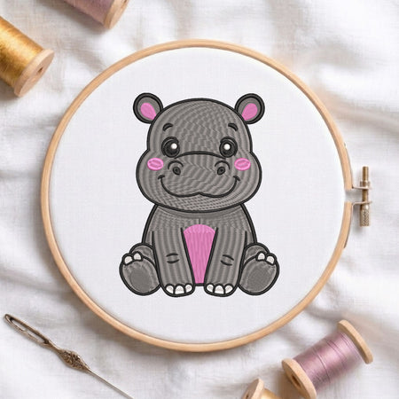A wooden embroidery hoop holding white fabric with a cartoon hippo design in gray, black, pink, and white. The hoop is photographed on a white fabric surface with spools of thread and a needle around it.