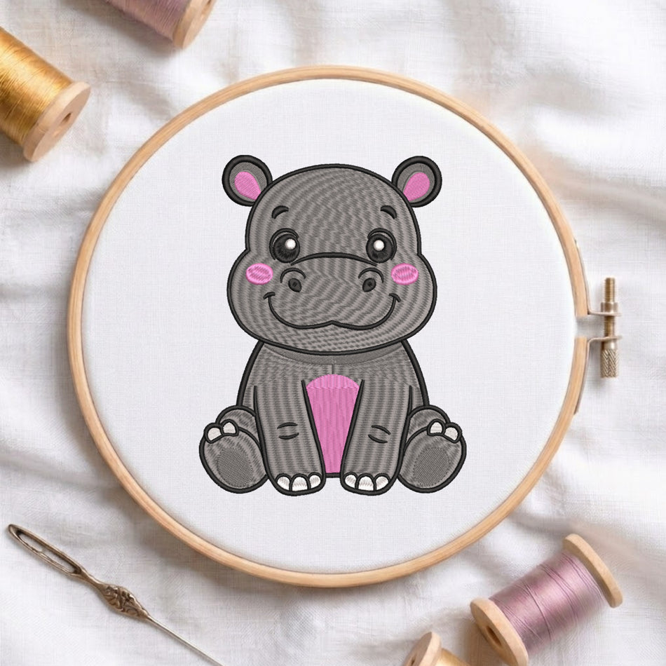 A wooden embroidery hoop holding white fabric with a cartoon hippo design in gray, black, pink, and white. The hoop is photographed on a white fabric surface with spools of thread and a needle around it.