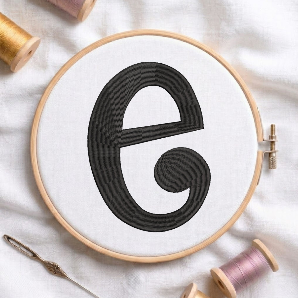 A wooden embroidery hoop holding white fabric with a large black stitched letter 'e' in a textured, striped stitch pattern. The hoop is styled on a white fabric background with spools of thread and a needle.