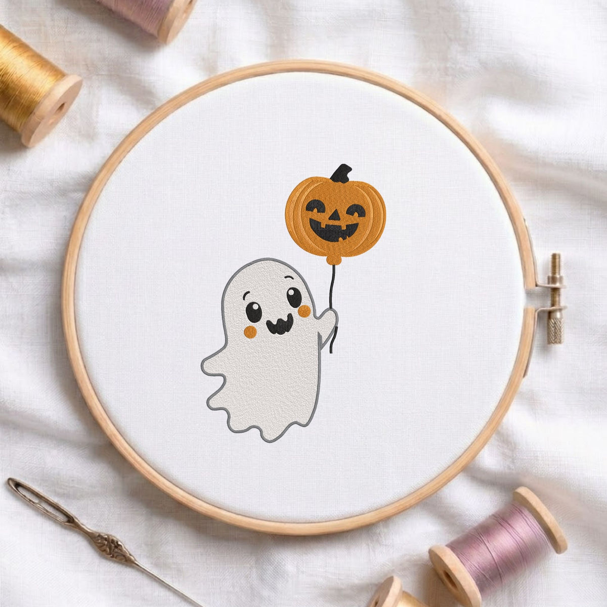 Cute ghost holding a jack-o-lantern pumpkin balloon machine embroidery design displayed in a wooden embroidery hoop on white fabric with thread spools and a needle   