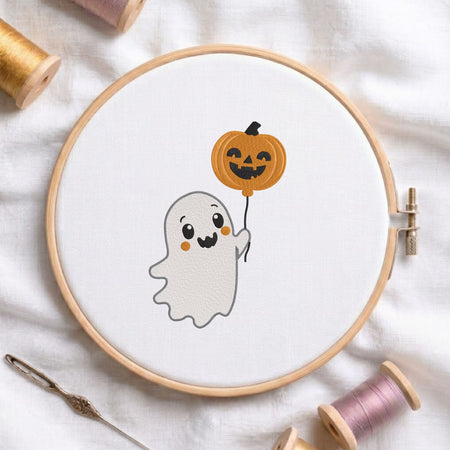 Cute ghost holding a jack-o-lantern pumpkin balloon machine embroidery design displayed in a wooden embroidery hoop on white fabric with thread spools and a needle   