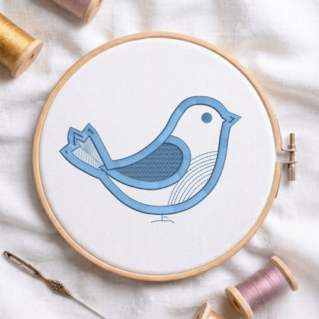 A round embroidery hoop holding white fabric with a stylized blue bird design stitched inside. The hoop is photographed on a white fabric surface with spools of thread and a needle around it.