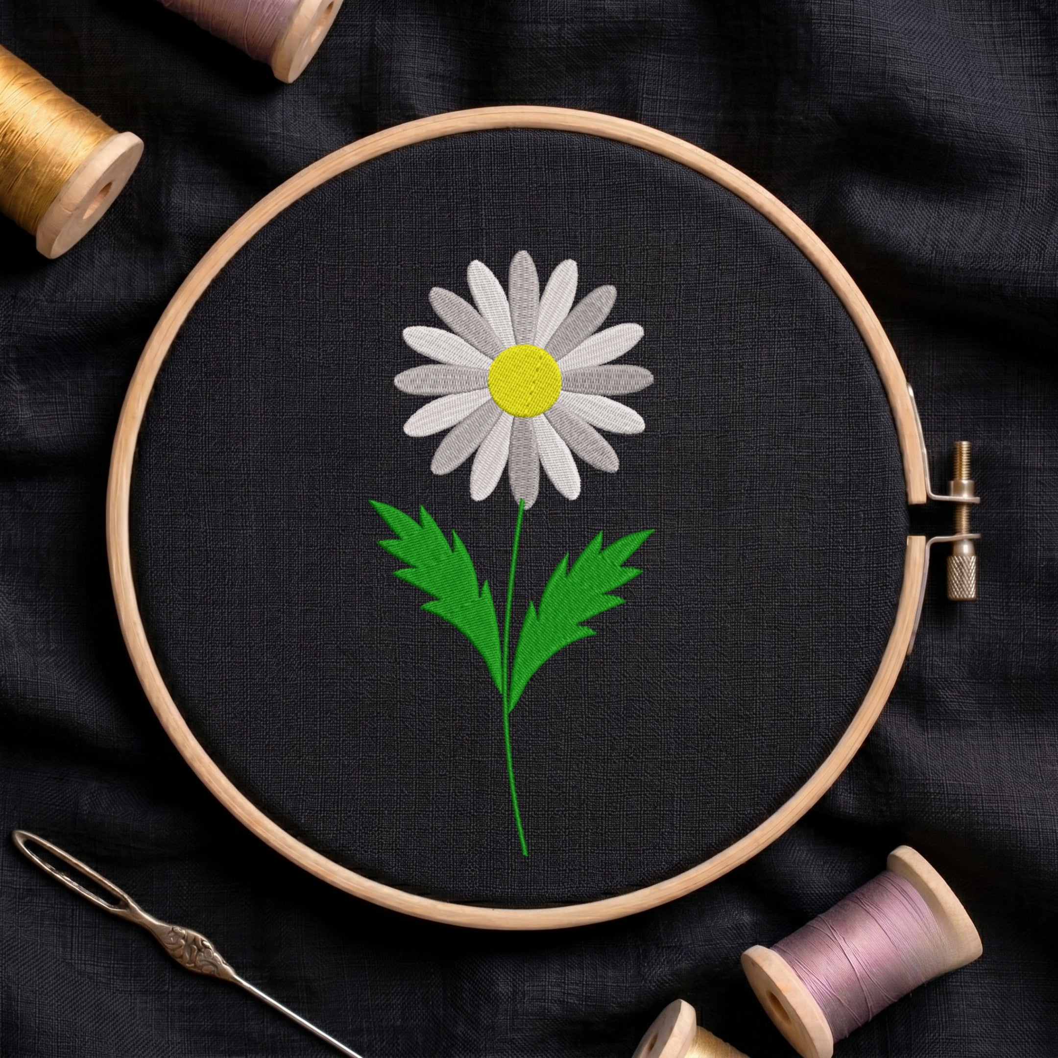 White daisy flower with a yellow center and green stem embroidered inside a round wooden hoop with thread spools against black fabric.