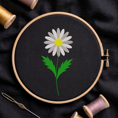 White daisy flower with a yellow center and green stem embroidered inside a round wooden hoop with thread spools against black fabric.