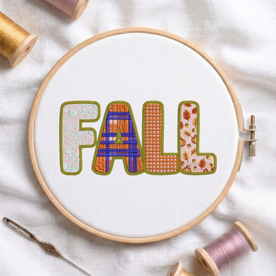 A circular embroidery hoop on white fabric displays the word “FALL” in decorative stitched letters. The hoop is surrounded by spools of thread and a needle, suggesting a crafting or sewing theme.