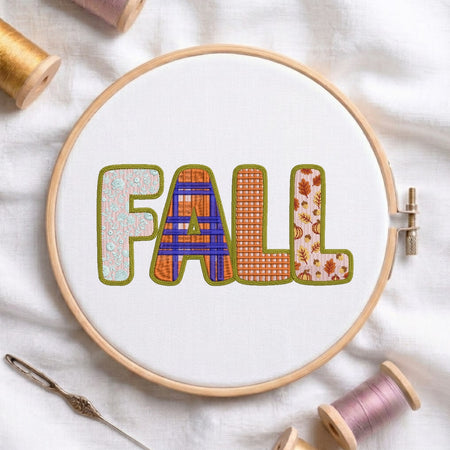 A circular embroidery hoop on white fabric displays the word “FALL” in decorative stitched letters. The hoop is surrounded by spools of thread and a needle, suggesting a crafting or sewing theme.