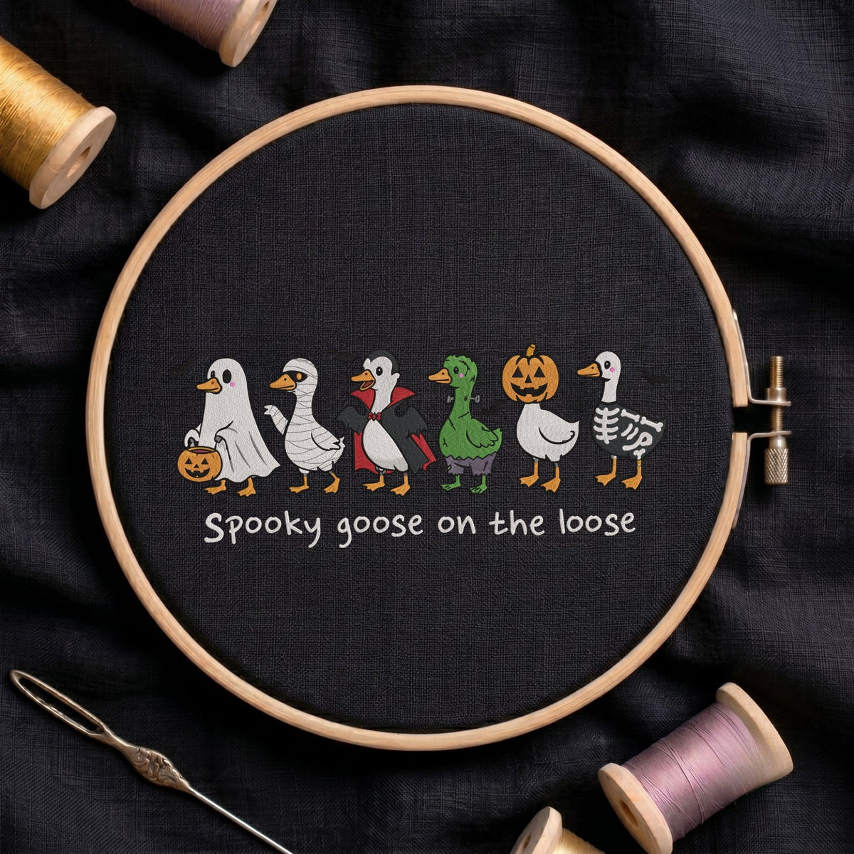 "Spooky goose on the loose" Halloween machine embroidery design featuring a line of cute geese dressed in spooky costumes including a ghost, mummy, vampire, Frankenstein monster, pumpkin head, and skeleton, shown in a wooden embroidery hoop on black fabric with thread spools and a needle   