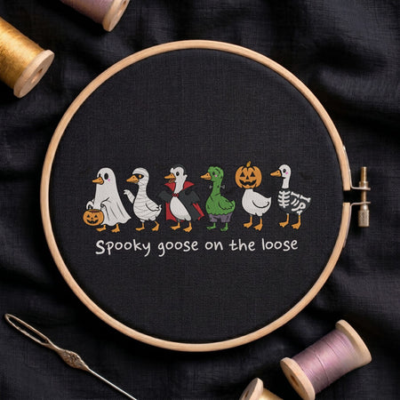 "Spooky goose on the loose" Halloween machine embroidery design featuring a line of cute geese dressed in spooky costumes including a ghost, mummy, vampire, Frankenstein monster, pumpkin head, and skeleton, shown in a wooden embroidery hoop on black fabric with thread spools and a needle   