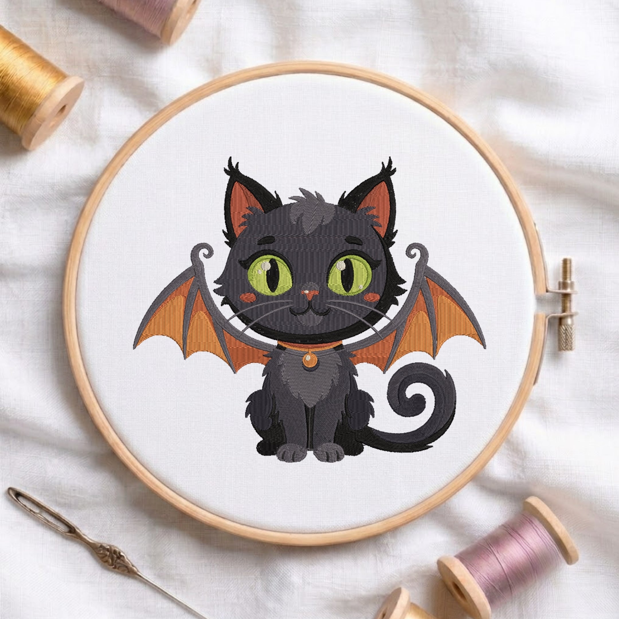 An embroidered black cat featuring green eyes, orange inner ears, and detailed bat wings, stitched inside a wooden hoop on white fabric with sewing threads.