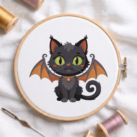 An embroidered black cat featuring green eyes, orange inner ears, and detailed bat wings, stitched inside a wooden hoop on white fabric with sewing threads.