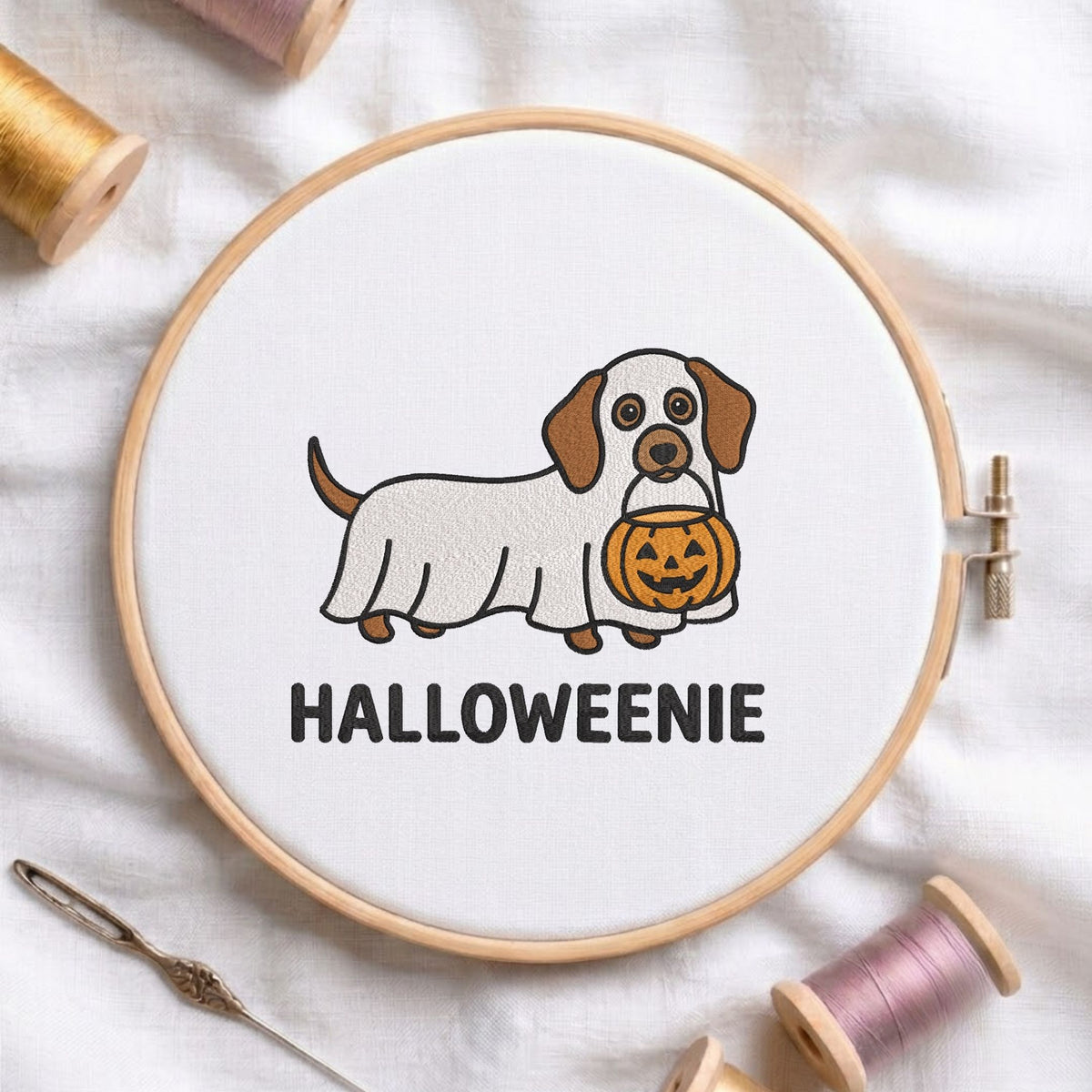 "Halloweenie" dachshund ghost holding a pumpkin machine embroidery design displayed in a wooden hoop on white fabric with thread spools and a needle