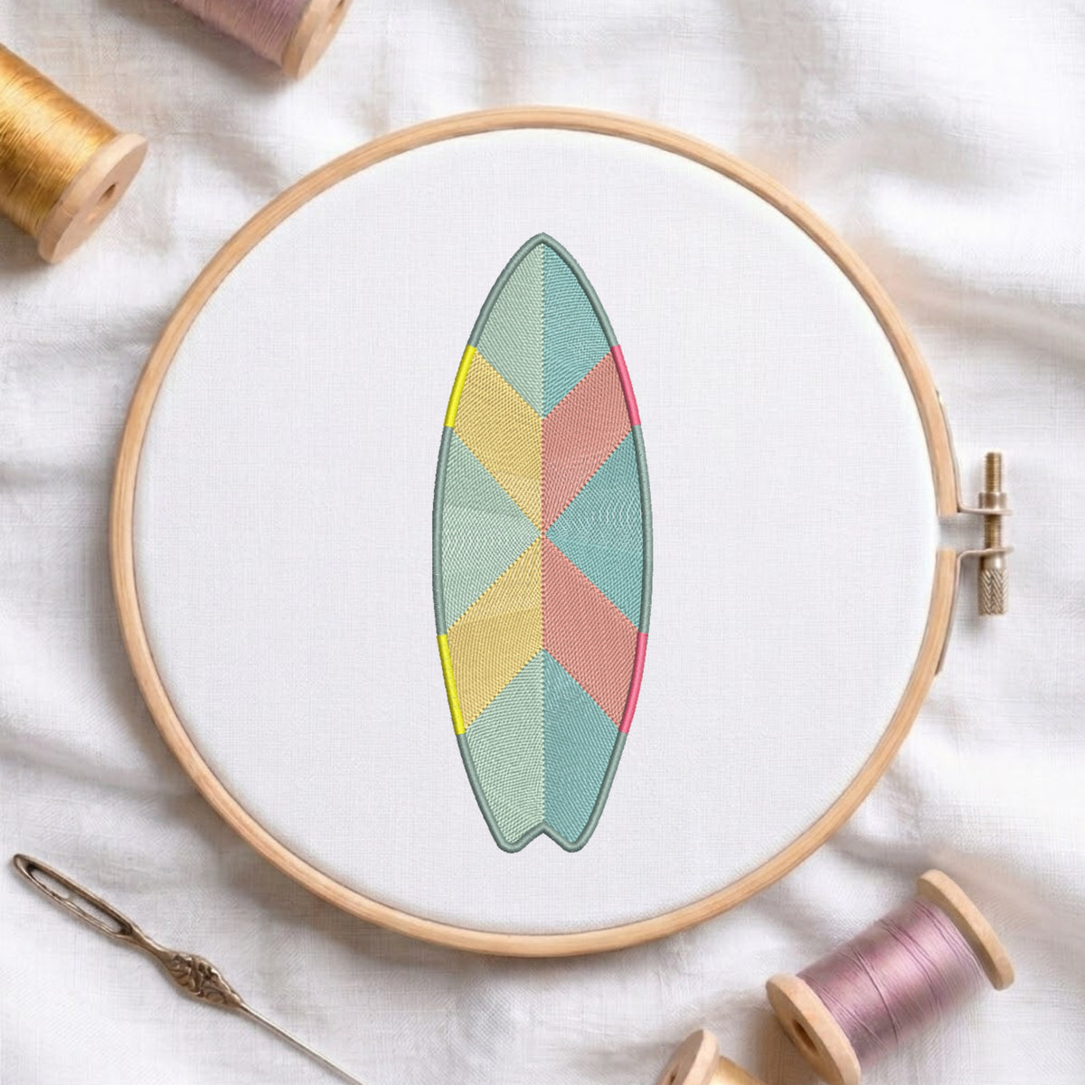 A wooden embroidery hoop holds white fabric with a surfboard-shaped geometric embroidery design in pastel teal, pink, yellow, and sage green. The hoop is styled on a white fabric background with spools of thread and a needle nearby.
