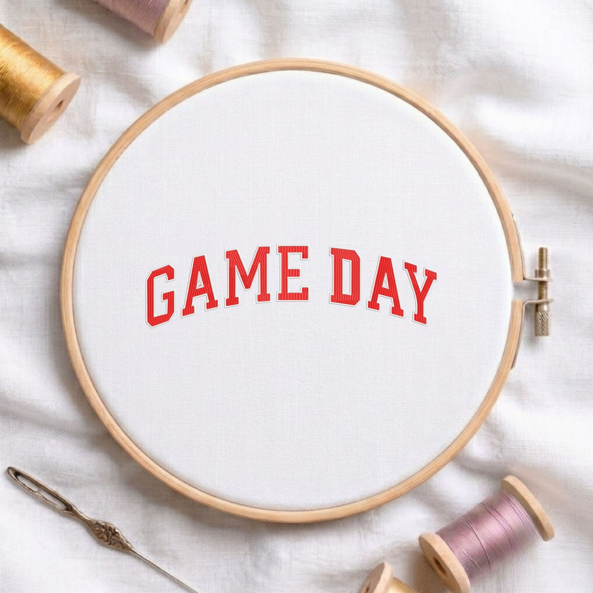 Red "GAME DAY" varsity athletic text machine embroidery design, displayed inside a round wooden hoop on white fabric with thread spools and a needle.