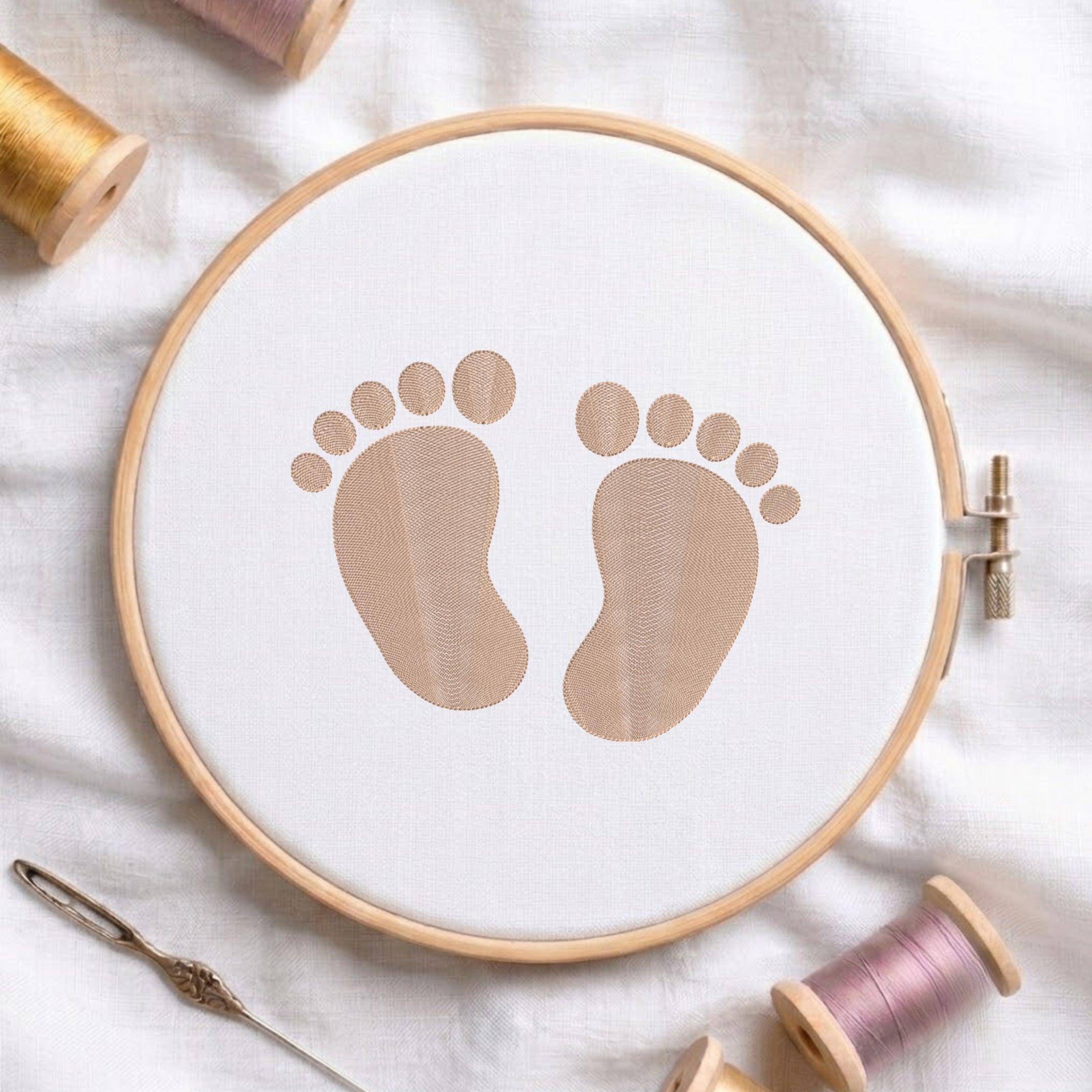 A circular embroidery hoop holding white fabric with a printed or stitched pair of baby footprints in a warm beige/tan color. The hoop is styled on a white fabric background with spools of thread and a needle around it.