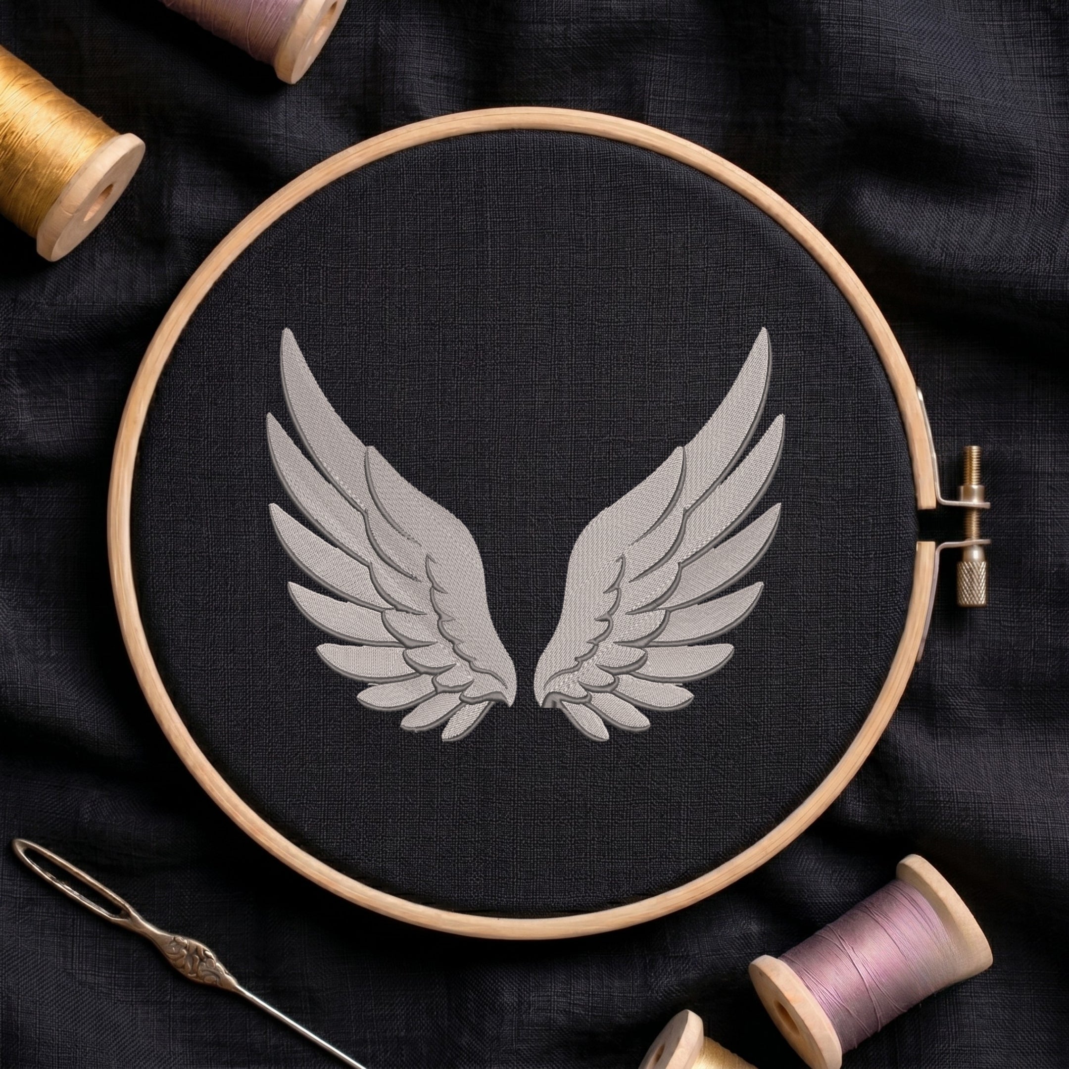 A wooden embroidery hoop holds black fabric with a stitched design of symmetrical gray wings. A metal needle lies at the lower left, and spools of thread in gold, pink, and purple are arranged around the hoop on a dark fabric background.