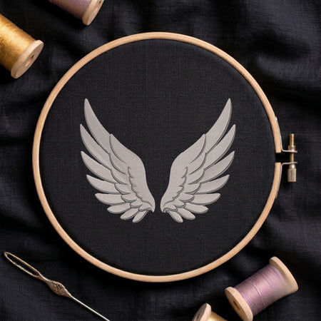 A wooden embroidery hoop holds black fabric with a stitched design of symmetrical gray wings. A metal needle lies at the lower left, and spools of thread in gold, pink, and purple are arranged around the hoop on a dark fabric background.