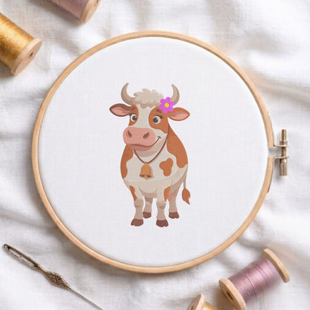 Cute cow with flower and bell machine embroidery design shown in a wooden embroidery hoop on white fabric with thread spools and a needle