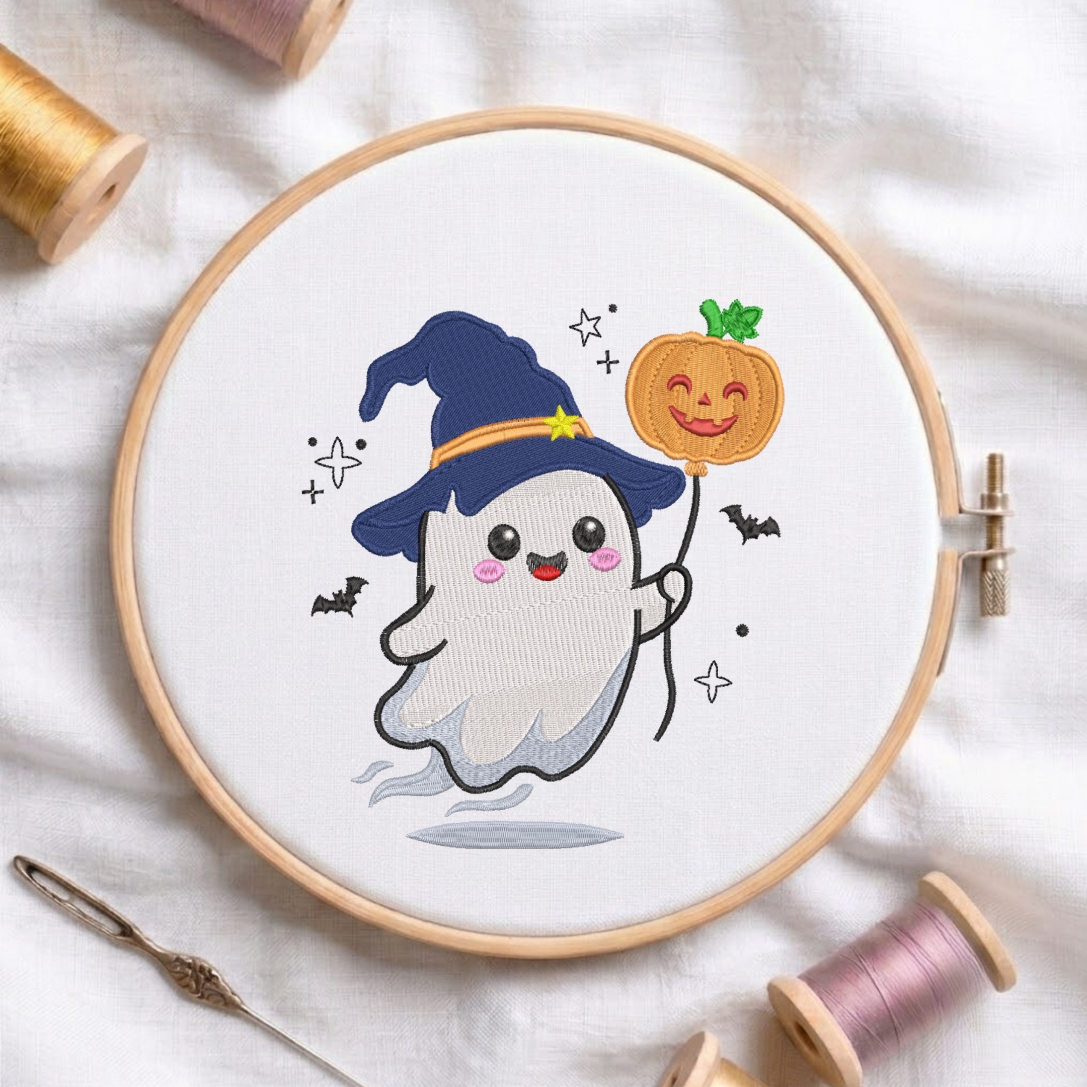 A circular embroidery hoop holding a white fabric with a cute cartoon ghost wearing a blue witch hat and holding a jack-o'-lantern, surrounded by spools of thread and a needle on a white fabric background.