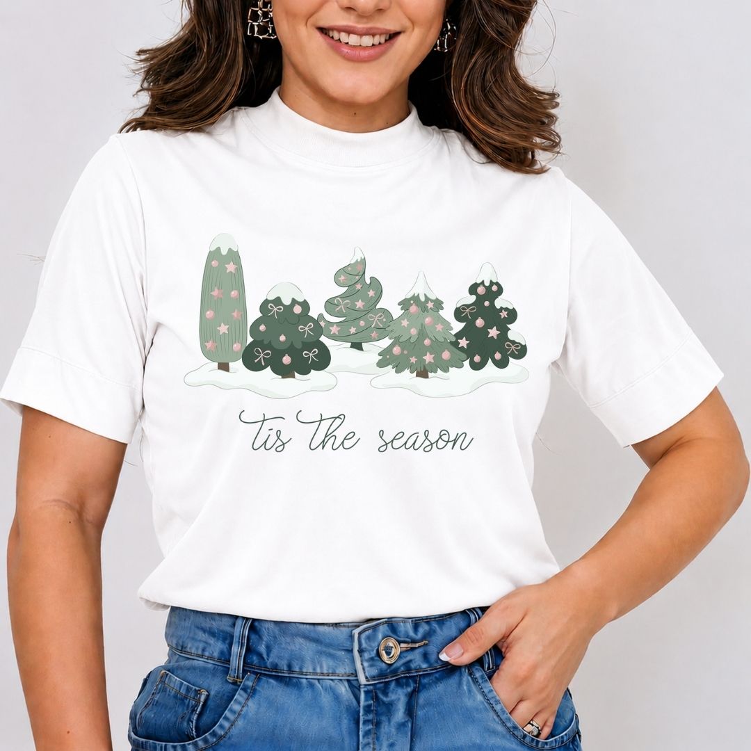 A woman wearing a white short-sleeve mock neck t-shirt with a graphic of five decorated Christmas trees and the text 'Tis the season' in script, paired with blue jeans.