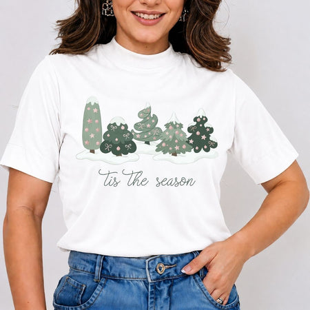 A woman wearing a white short-sleeve mock neck t-shirt with a graphic of five decorated Christmas trees and the text 'Tis the season' in script, paired with blue jeans.