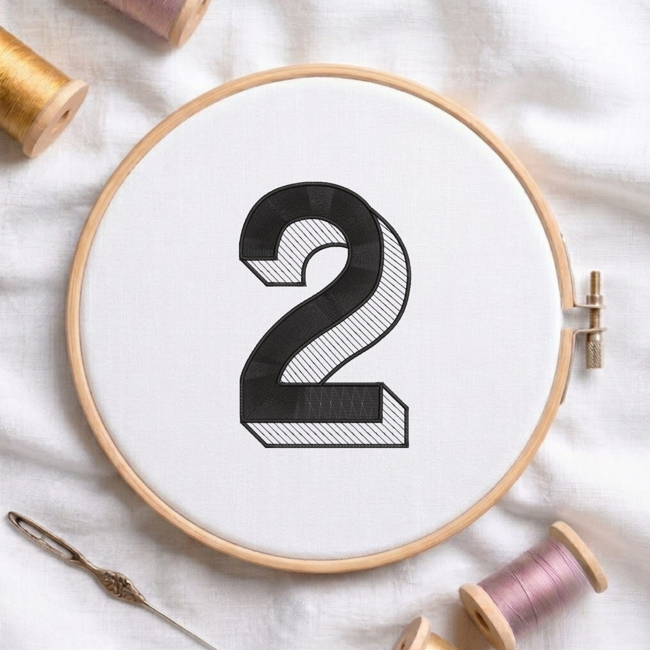 A wooden embroidery hoop holding white fabric with a large black number 2 stitched in the center, surrounded by spools of thread and a needle on a white fabric background.