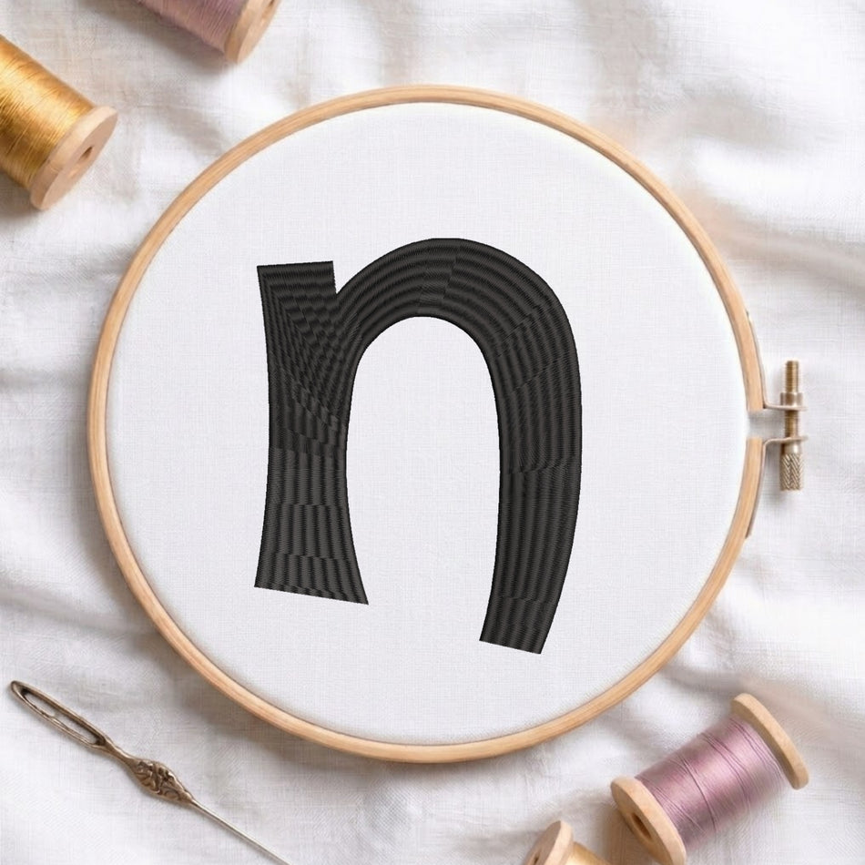 A wooden embroidery hoop holding white fabric with a large black letter 'n' stitched in the center. A metal needle lies nearby on white fabric, with spools of thread partially visible around the edges.