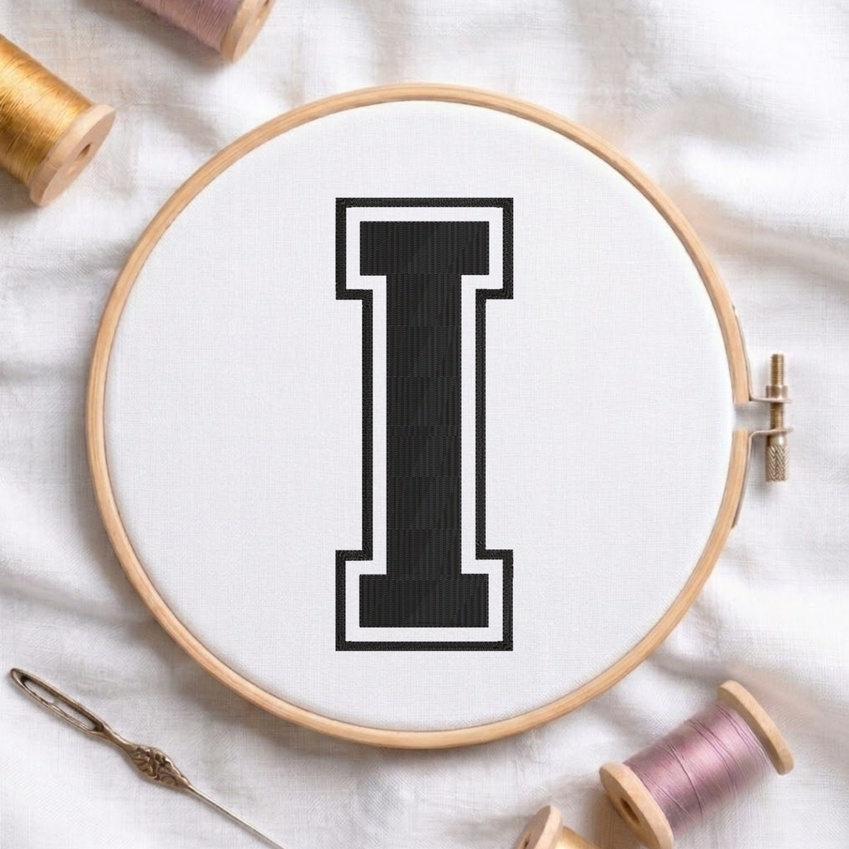A wooden embroidery hoop holding white fabric with a large black letter 'I' stitched in the center, surrounded by spools of thread and a needle on a white fabric background.