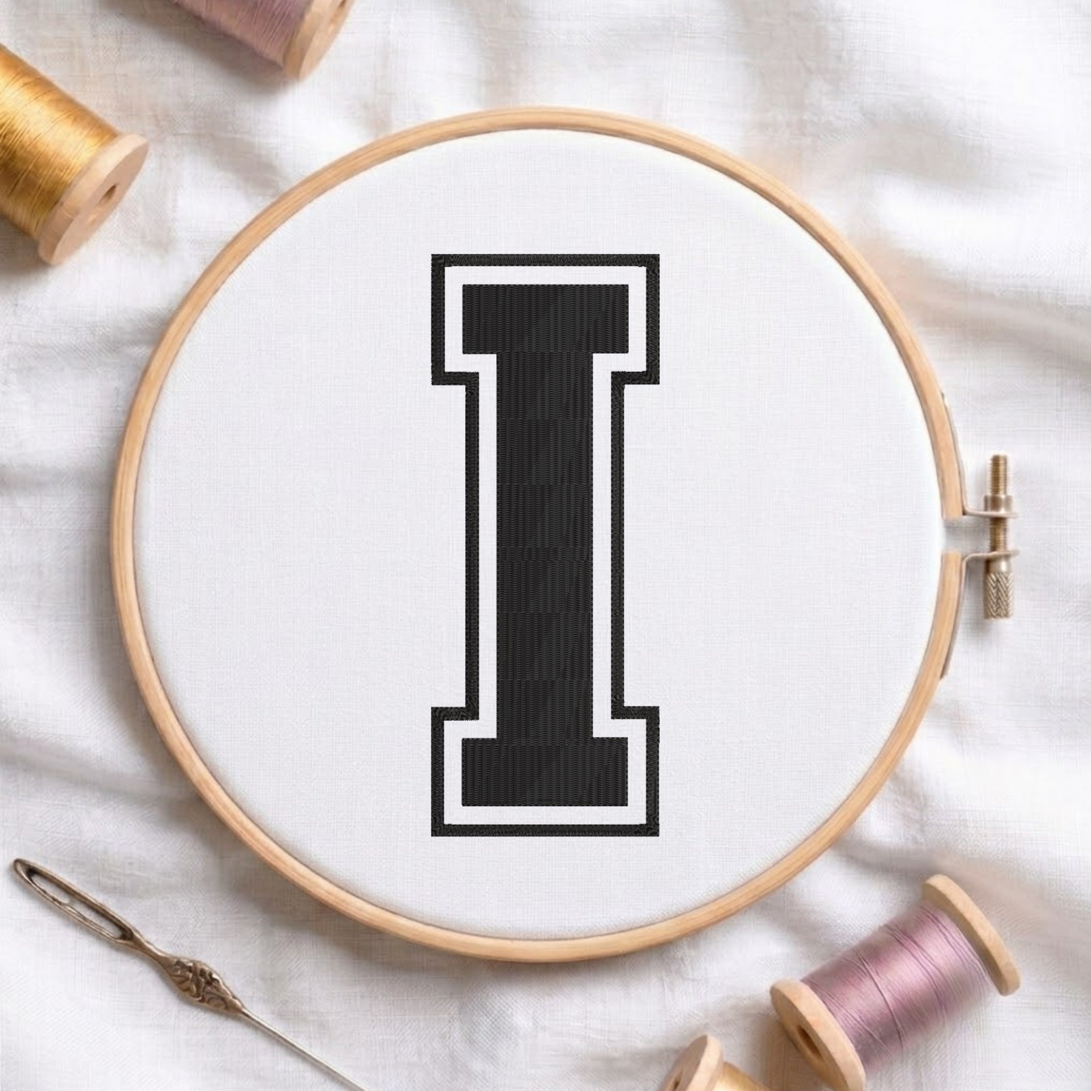 A wooden embroidery hoop holding white fabric with a large black letter 'I' stitched in the center, surrounded by spools of thread and a needle on a white fabric background.