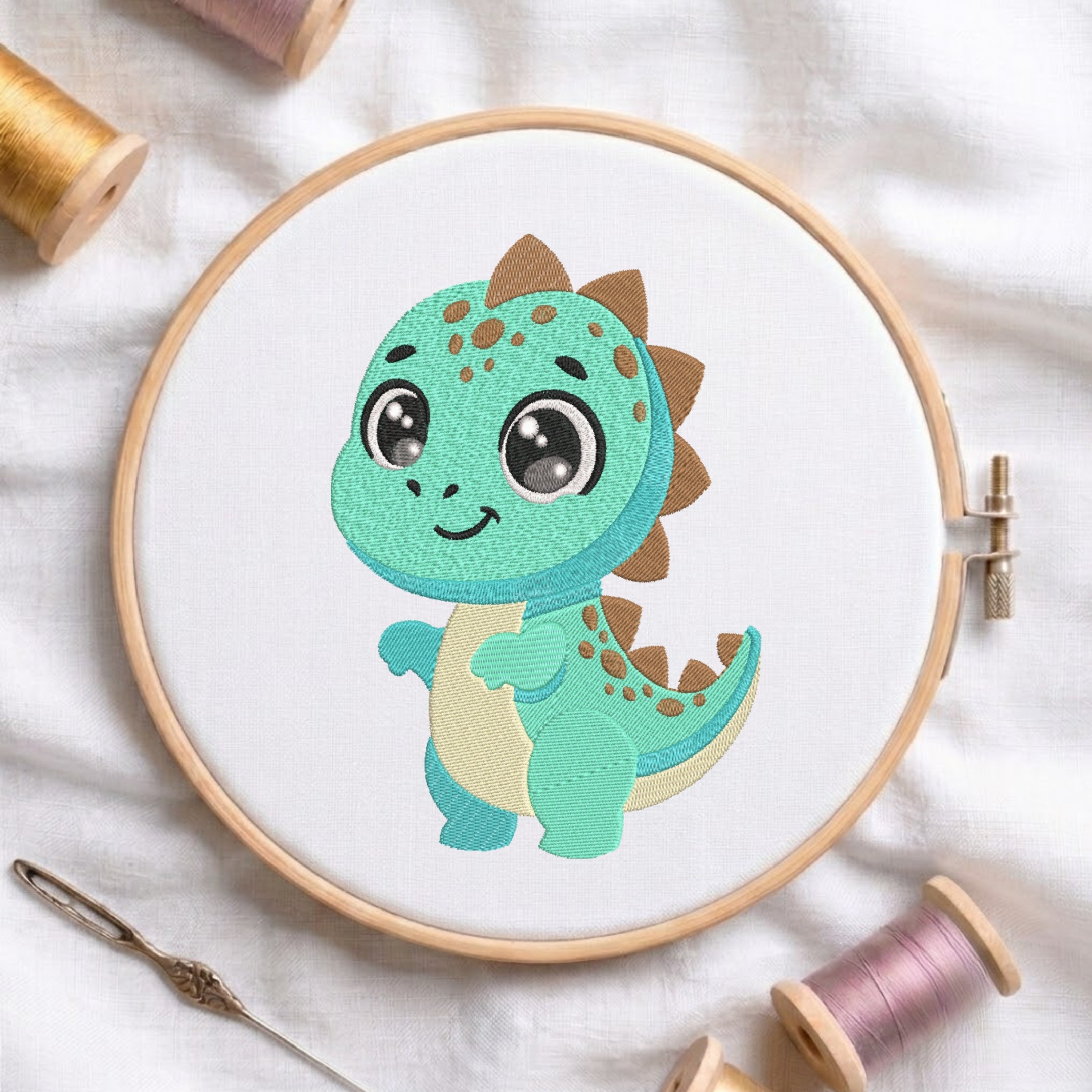 A wooden embroidery hoop holds a white fabric panel featuring a cute cartoon dinosaur design in mint green, teal, cream, and brown. The hoop is styled on a white fabric background with spools of thread and a needle.