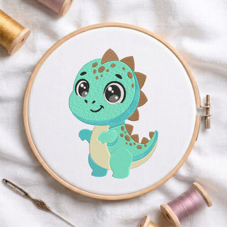 A wooden embroidery hoop holds a white fabric panel featuring a cute cartoon dinosaur design in mint green, teal, cream, and brown. The hoop is styled on a white fabric background with spools of thread and a needle.