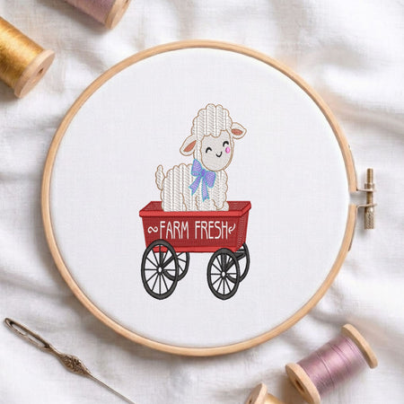  Cute fluffy sheep with a purple bow sitting inside a red "Farm Fresh" wagon machine embroidery design, displayed inside a round wooden hoop on white fabric with thread spools and a needle.   