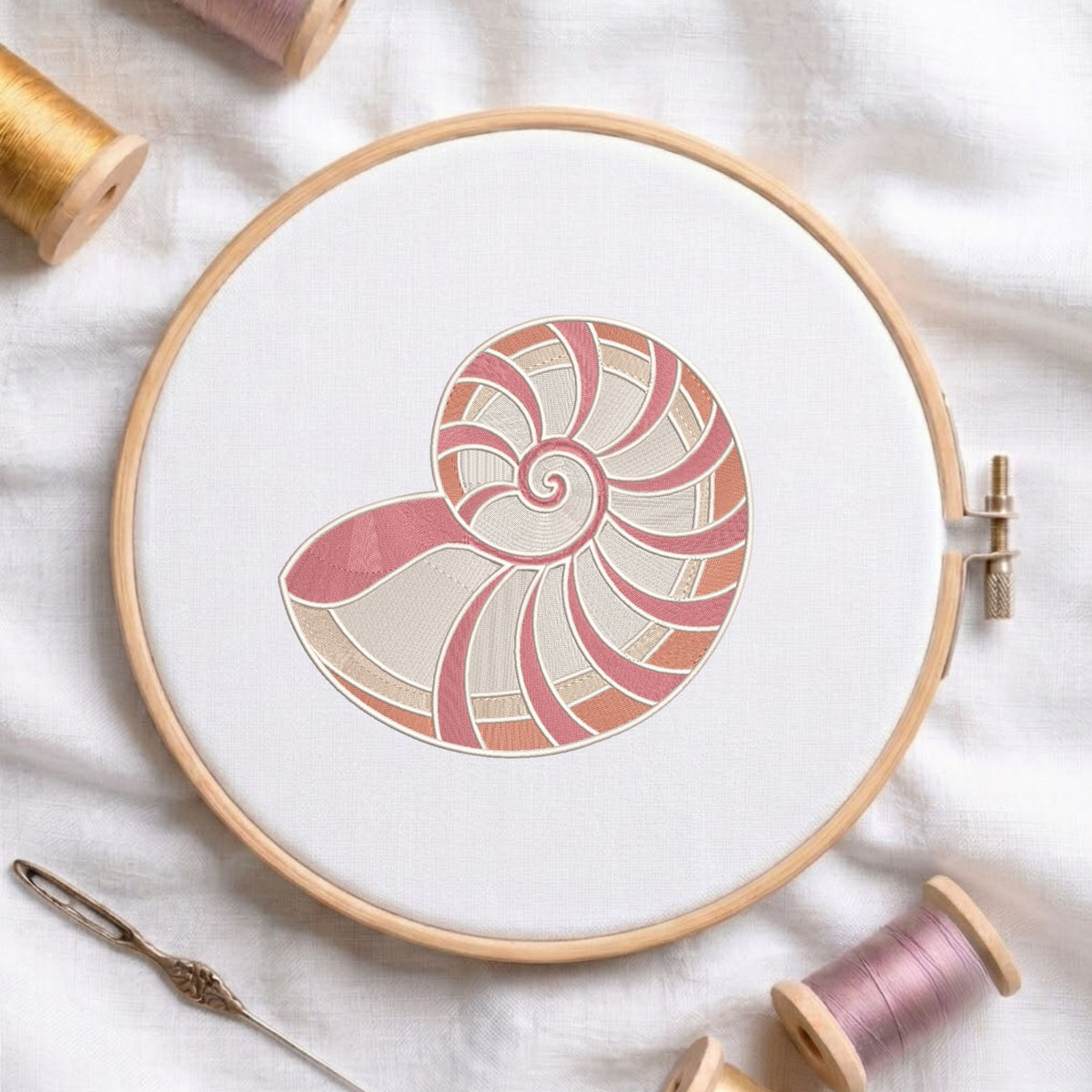 A wooden embroidery hoop holds white fabric with a stylized nautilus shell design in pink, peach, and cream. Spools of thread and a needle are arranged around it on a white fabric background.