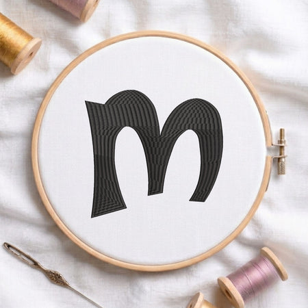 A wooden embroidery hoop holding white fabric with a large black embroidered letter 'm' in the center. A silver needle lies nearby on a white fabric background, with spools of thread partially visible around the hoop.