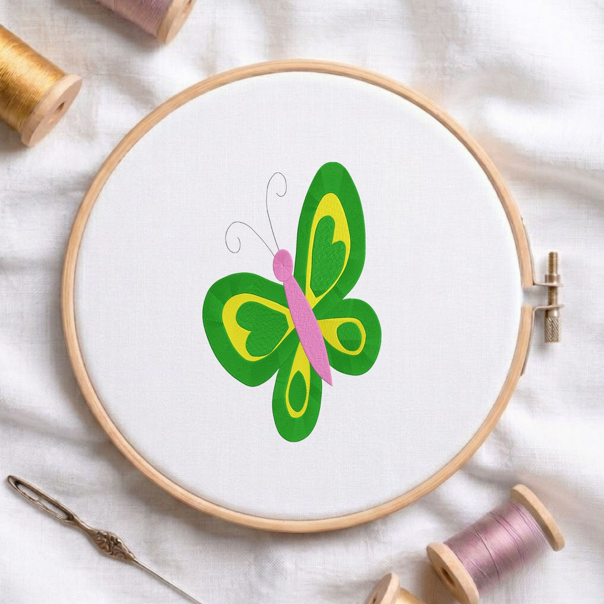 Green and yellow butterfly machine embroidery design displayed in a wooden embroidery hoop on white fabric with thread spools and a needle   