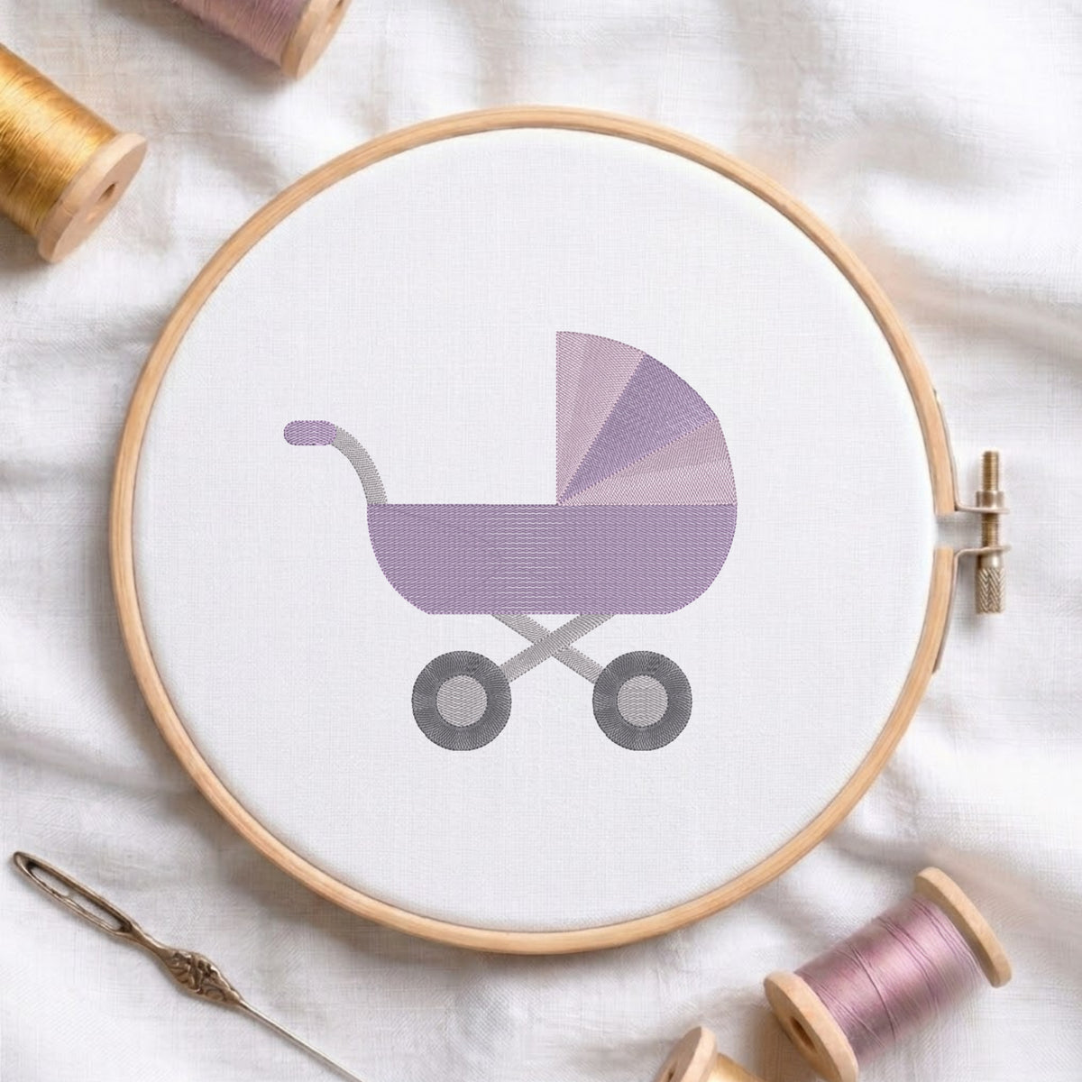 A circular embroidery hoop holding white fabric with a stylized lavender baby stroller design, surrounded by spools of thread and a needle on a white fabric background.