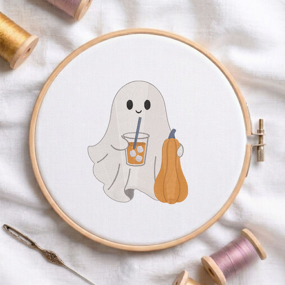 Cute ghost drinking iced coffee and holding a pumpkin machine embroidery design displayed in a wooden hoop on white fabric with thread spools and a needle