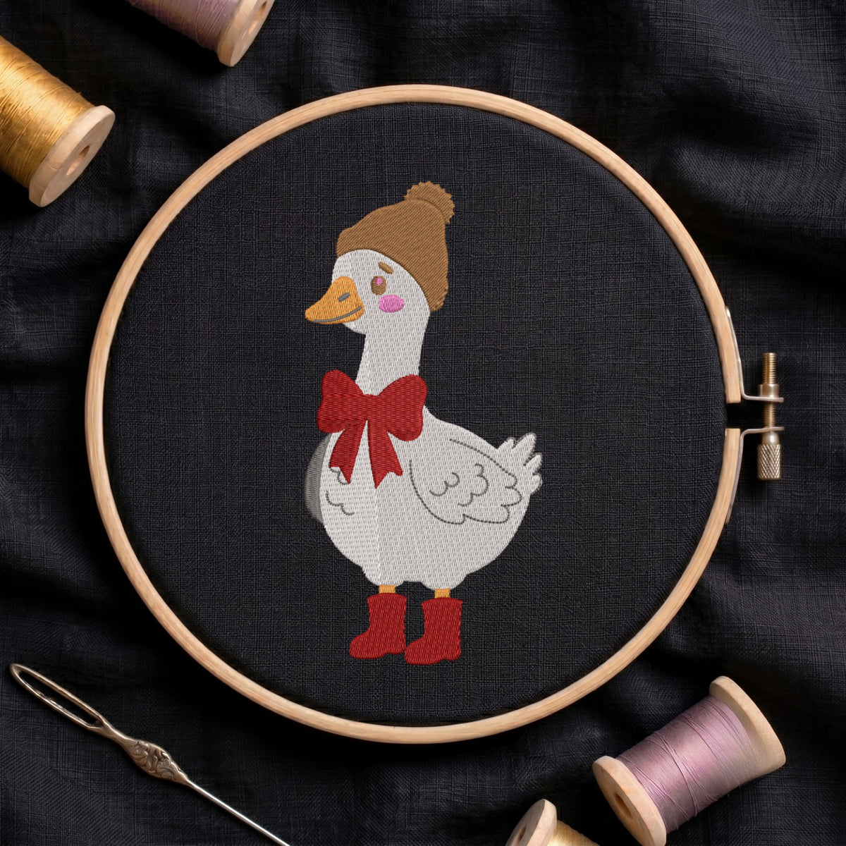A circular embroidery hoop holds a white goose stitched on black fabric, wearing a brown knit hat, a red bow, and red boots. A needle lies at the lower left, and spools of thread are partially visible around the hoop on a dark fabric background.