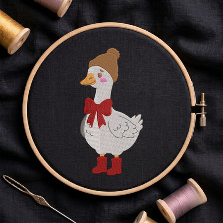 A circular embroidery hoop holds a white goose stitched on black fabric, wearing a brown knit hat, a red bow, and red boots. A needle lies at the lower left, and spools of thread are partially visible around the hoop on a dark fabric background.