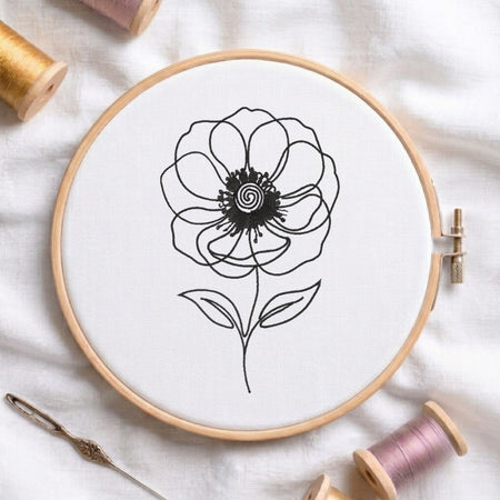 A wooden embroidery hoop holding white fabric with a black line-art flower design, surrounded by spools of thread and a needle on a white fabric background.