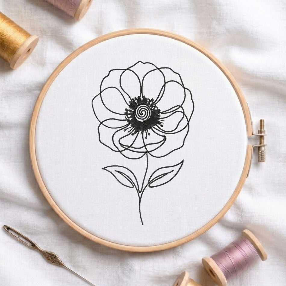 A wooden embroidery hoop holding white fabric with a black line-art flower design, surrounded by spools of thread and a needle on a white fabric background.
