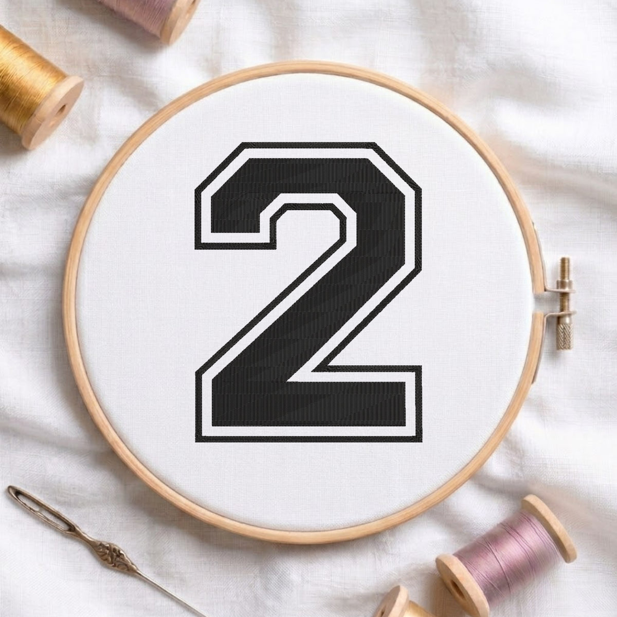 A wooden embroidery hoop holding white fabric with a large black number 2 outlined in white, styled on a white fabric background with spools of thread and a needle.