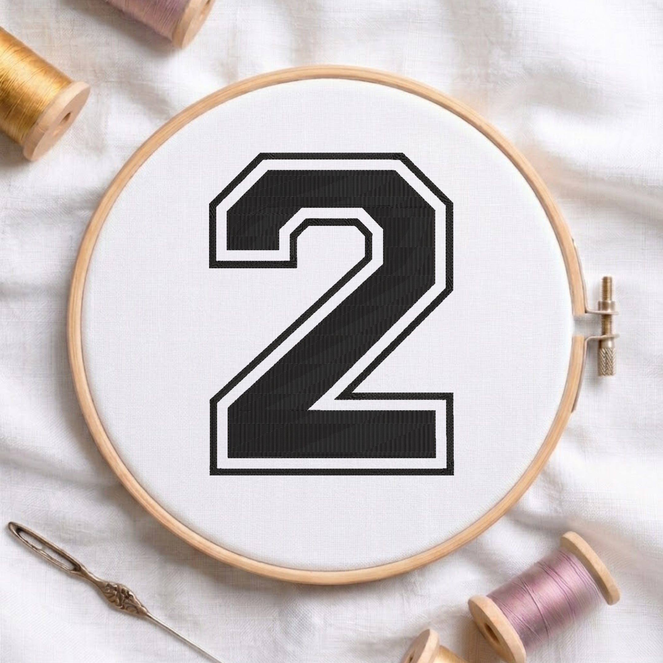 A wooden embroidery hoop holding white fabric with a large black number 2 outlined in white, styled on a white fabric background with spools of thread and a needle.