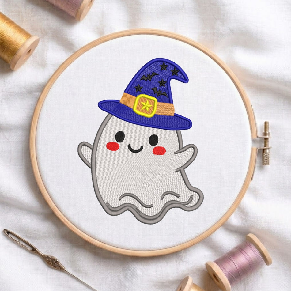 A circular embroidery hoop holding white fabric with a cute embroidered ghost wearing a purple wizard hat with black stars and a yellow band. The hoop is styled on a white fabric background with spools of thread and a needle.