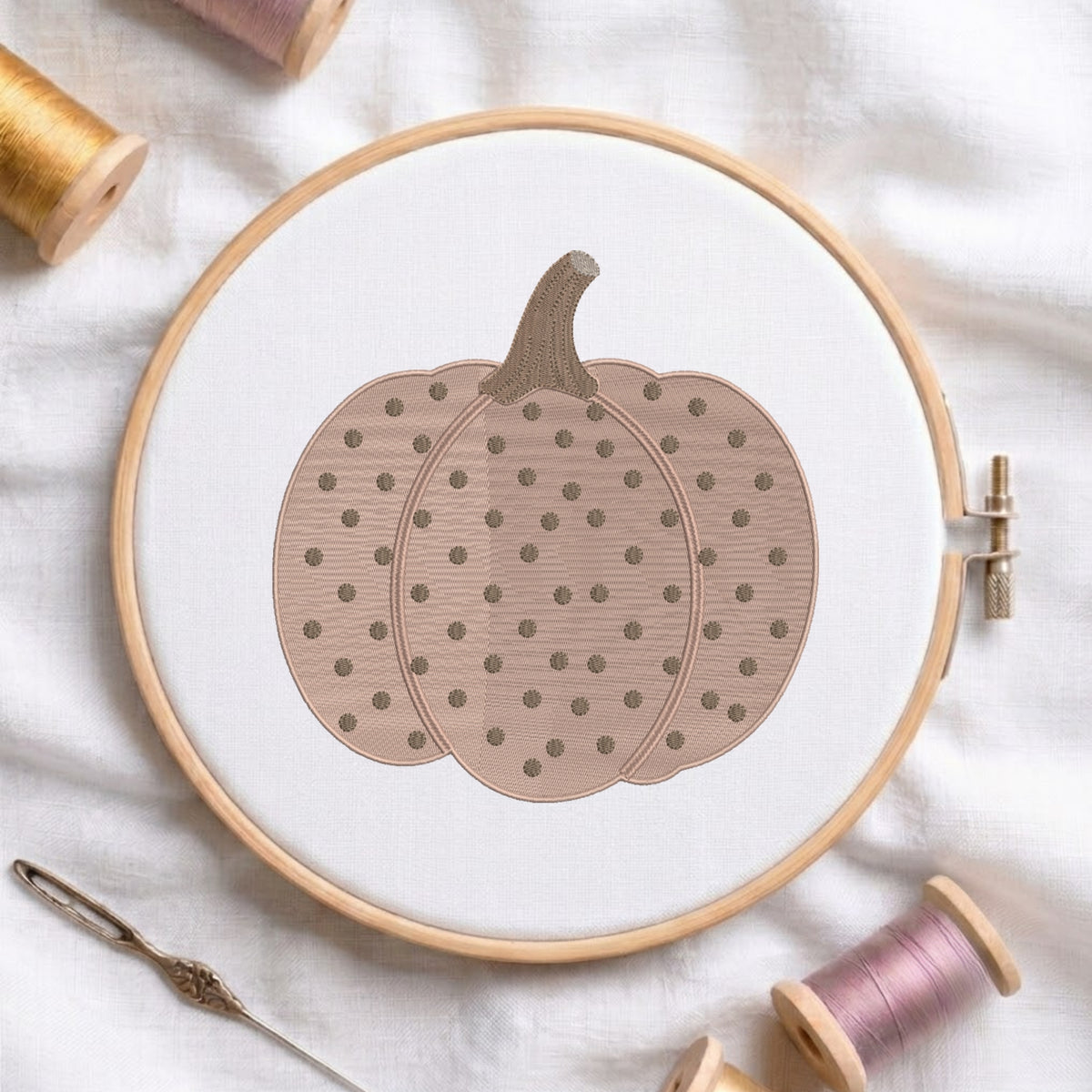 A circular embroidery hoop holding white fabric with a stitched pumpkin design in muted taupe and brown, surrounded by spools of thread and a needle on a white fabric background.