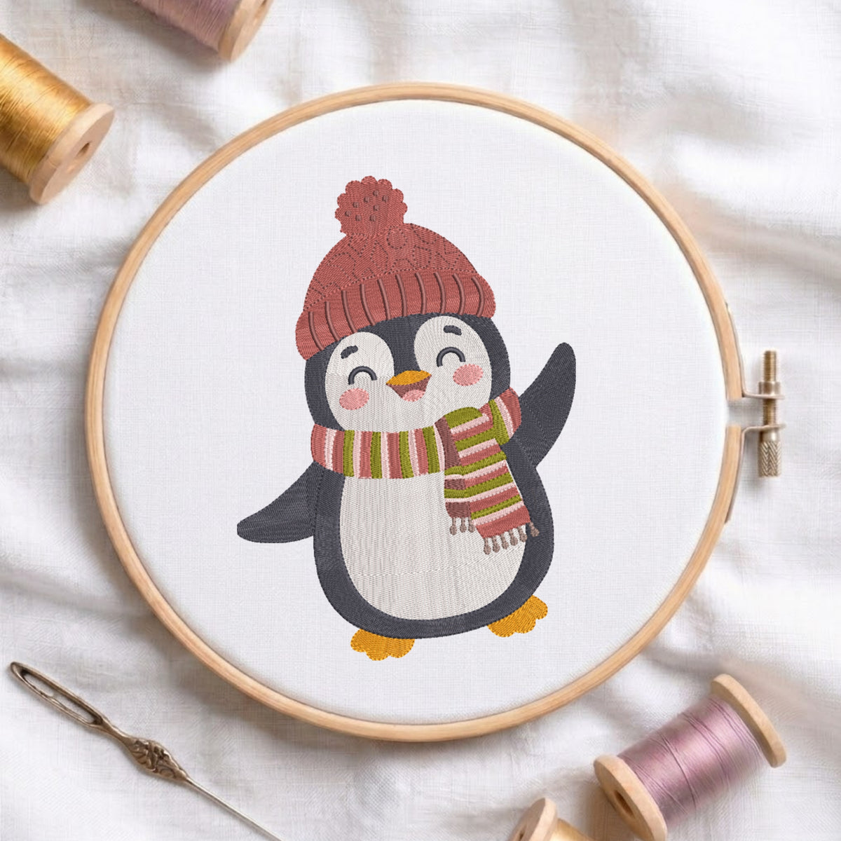 A circular embroidery hoop holding white fabric with a stitched penguin design wearing a red knit hat and a striped red, green, and yellow scarf. The hoop is photographed on a white fabric surface with spools of thread and a needle around it.