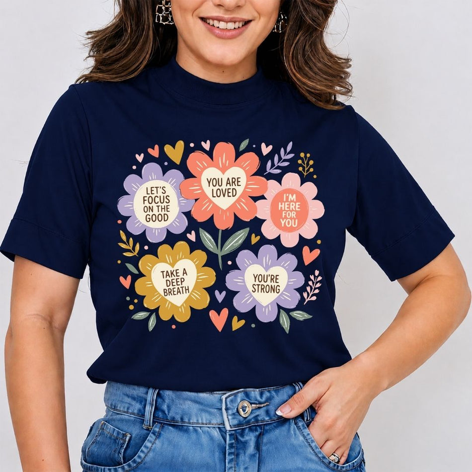 A woman wearing a navy blue graphic t-shirt with colorful floral designs and motivational quotes, paired with blue jeans, against a light gray background.