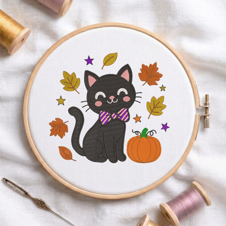 A circular embroidery hoop on white fabric, featuring a black cat wearing a purple striped bow tie, surrounded by autumn leaves, stars, and an orange pumpkin. Spools of thread and a needle are arranged around the hoop.