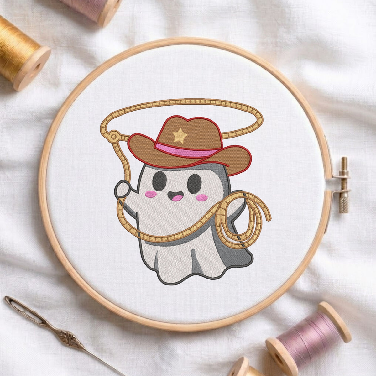 A circular embroidery hoop displays a cute cartoon ghost cowboy design with a brown hat, pink band, and rope lasso. The hoop is styled on a white fabric background with spools of thread and a needle.