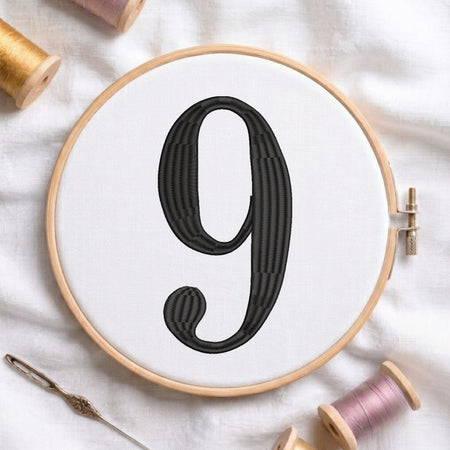 A wooden embroidery hoop holds white fabric with a large black stitched number 9. A metal embroidery needle rests near the bottom left, and spools of thread are arranged around the hoop on a white fabric background.