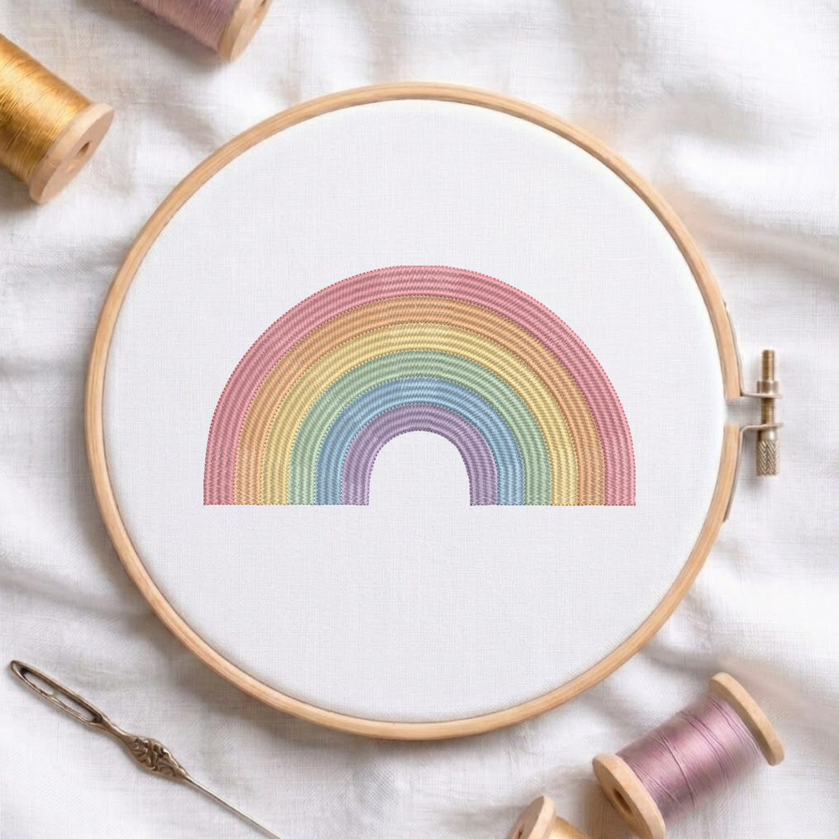 A wooden embroidery hoop holding white fabric with a pastel rainbow stitched design, surrounded by spools of thread and a needle on a white fabric background.